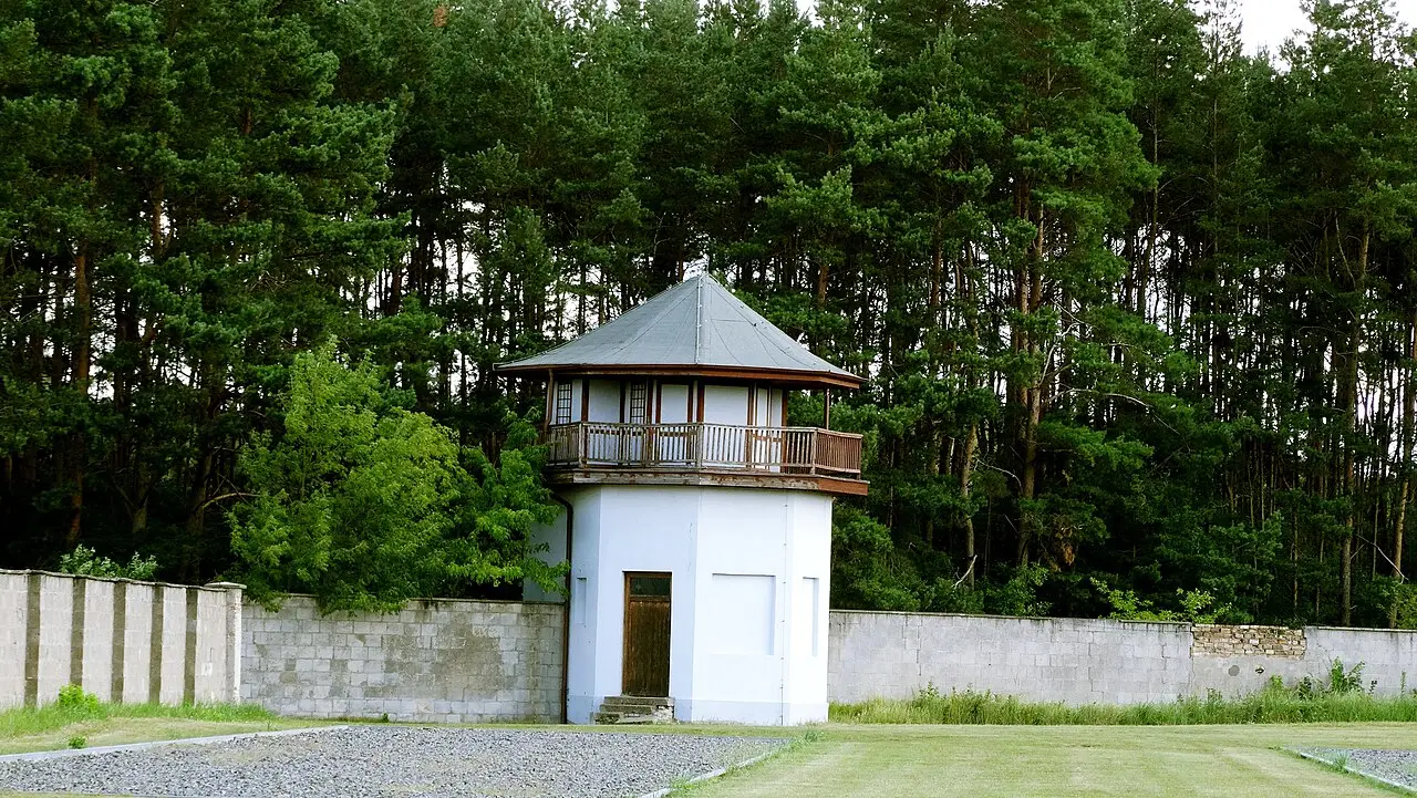 Torre de guarda branca junto ao muro de blocos de concreto do antigo campo de Sachsenhausen, com pinheiros altos ao fundo.