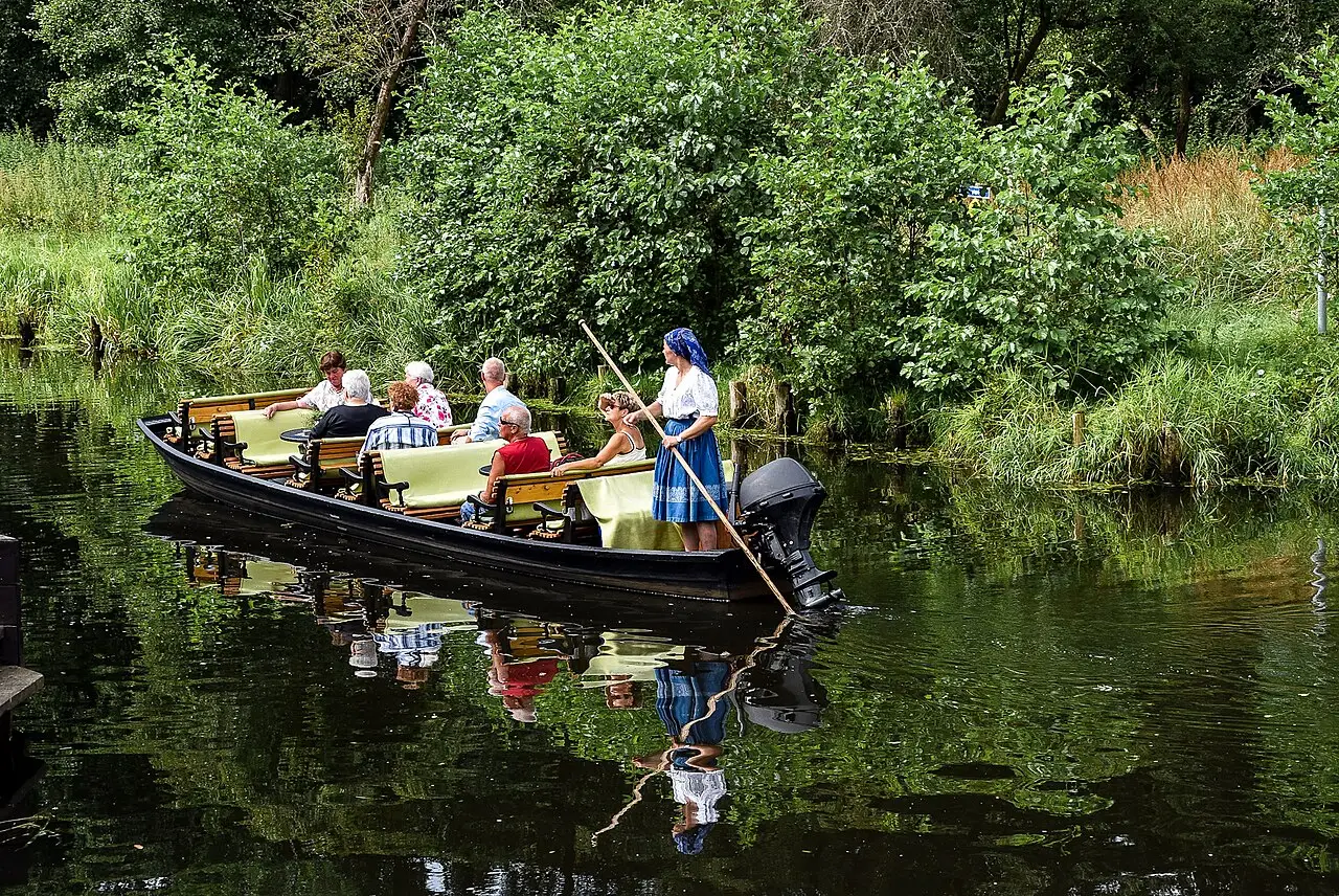 Barco de passeio em um canal do Spreewald, com passageiros sentados e uma condutora de saia azul com uma vara, entre vegetação verde.