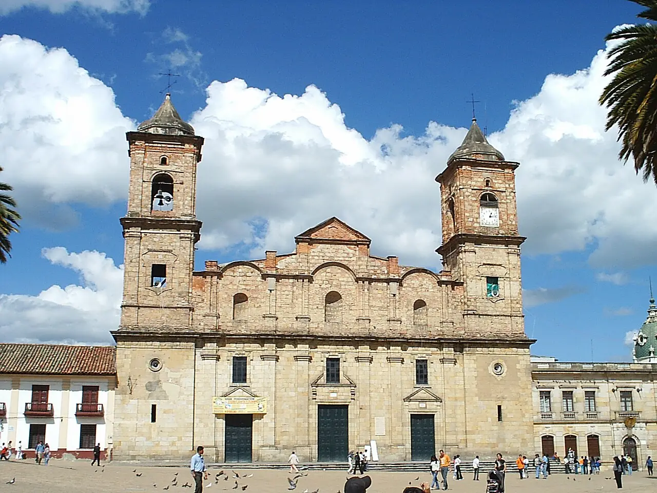 Fachada de pedra de uma igreja com duas torres na praça central de Zipaquirá, com pessoas e pombos na praça e nuvens no céu.