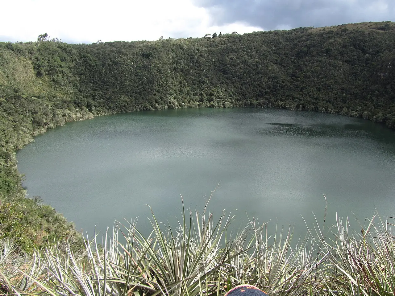 Laguna de Guatavita vista de cima, um lago redondo de água verde-acinzentada cercado por encostas cobertas de mata densa.