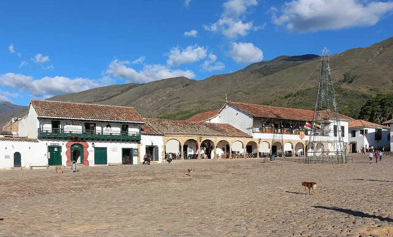 Plaza Mayor de Villa de Leyva, um grande espaço de pedras irregulares cercado de casas brancas de telhado de barro, com montanhas ao fundo.