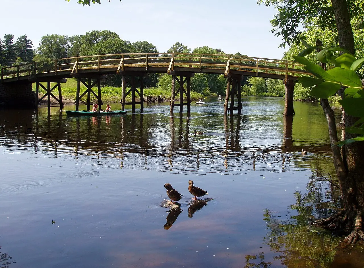 Puente arqueado de madera sobre el río Concord, entre orillas verdes con árboles, bajo un cielo azul.