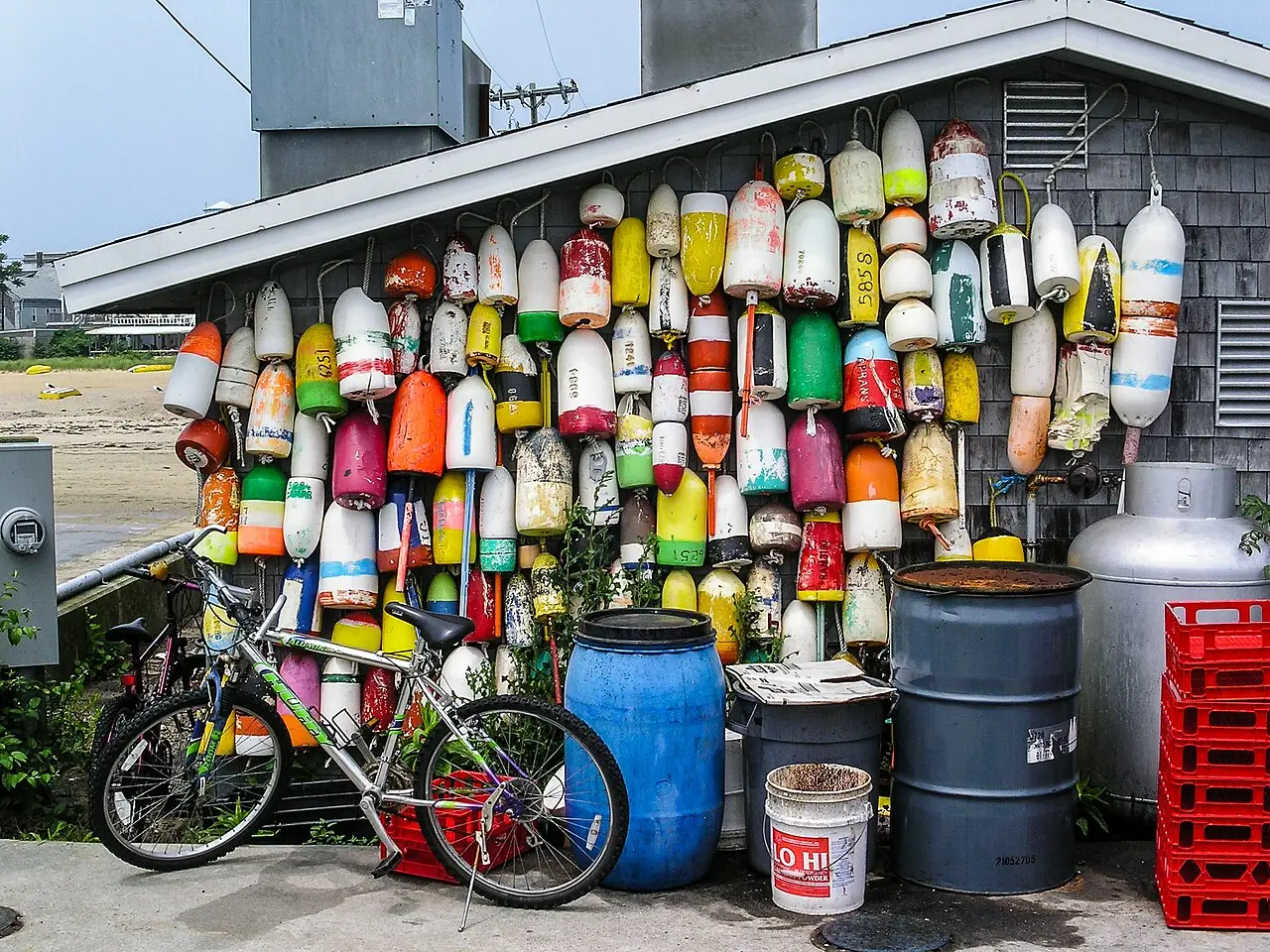 Parede de uma cabana de pescador em Provincetown coberta de boias coloridas, com uma bicicleta, tambores e uma faixa de praia ao lado.