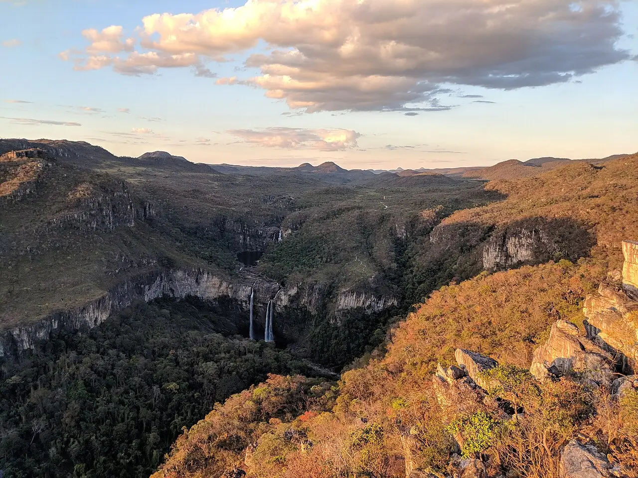 Vista do Mirante da Janela no Parque Nacional da Chapada dos Veadeiros, com um vale de vegetação e duas quedas d’água ao fundo ao entardecer.