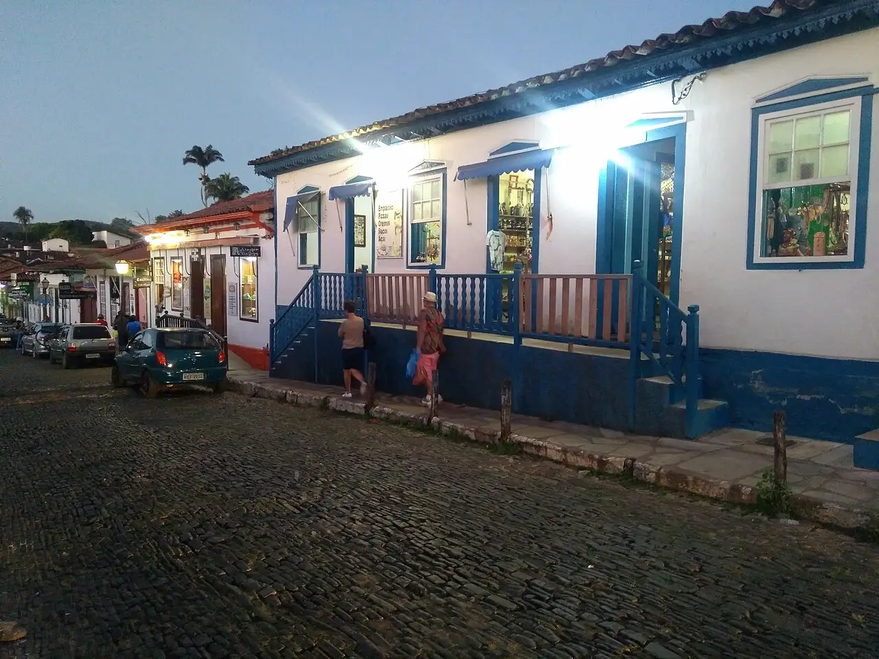 Rua de paralelepípedos em Pirenópolis ao anoitecer, com casarão colonial branco de detalhes azuis, lojas iluminadas e pedestres na calçada.