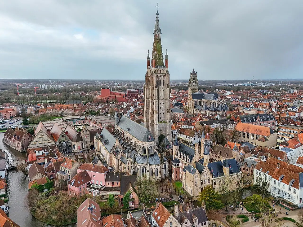 Vista aérea de Bruges com a alta torre da Igreja de Nossa Senhora no centro, telhados vermelhos, um canal à esquerda e céu nublado.