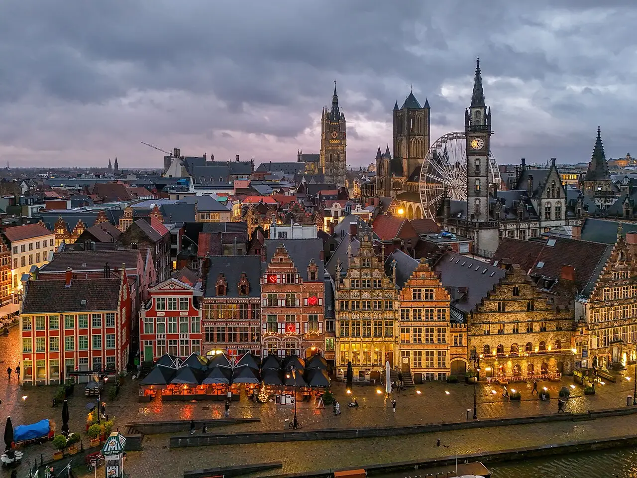 Vista aérea de Gante ao entardecer, com as casas de fachada em degraus da Graslei iluminadas, torres de igrejas e o campanário ao fundo.