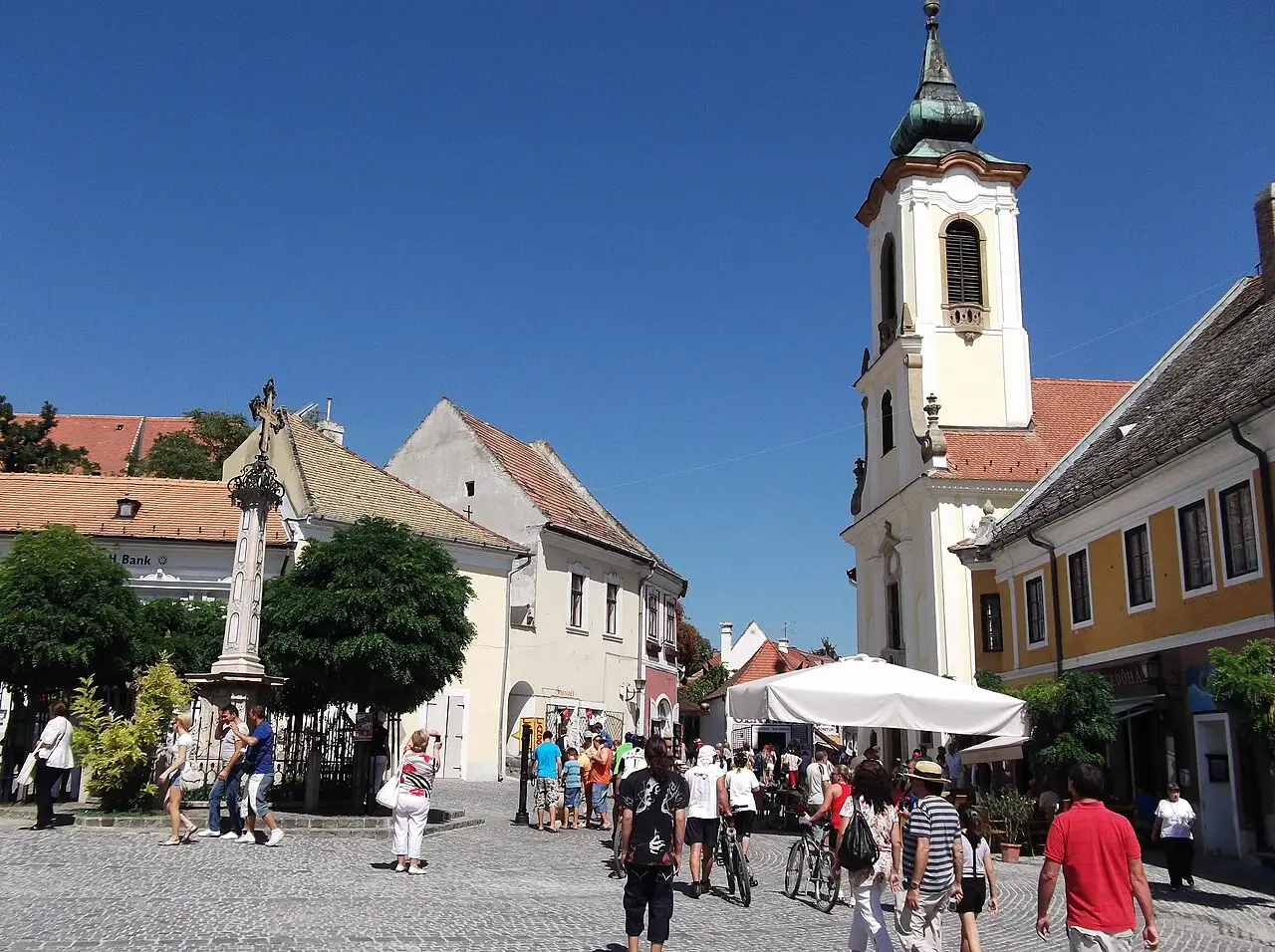 Praça de Szentendre com igreja de torre barroca branca, casas de telhado vermelho, uma cruz de pedra e muitos visitantes a pé e de bicicleta.