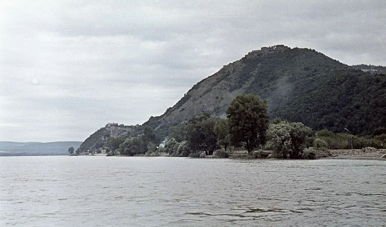 Fotografia antiga colorida do Danúbio em Visegrád, com um morro arborizado e a cidadela em ruínas no alto, visto da água.