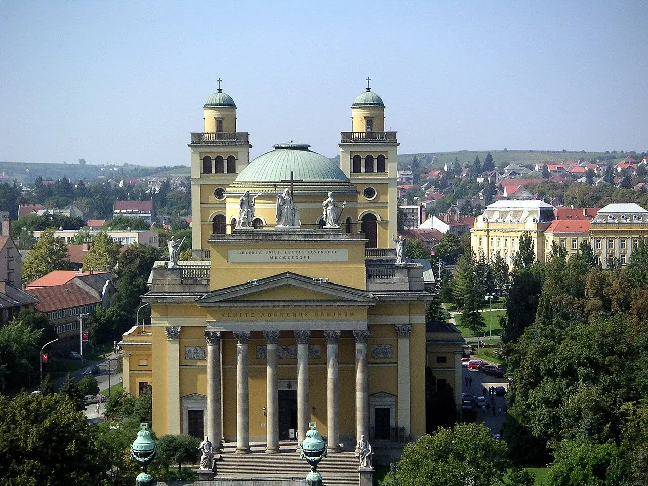 Fachada amarela da Basílica de Eger, com pórtico de colunas, duas torres com cúpulas verdes, uma cúpula central e estátuas no alto.