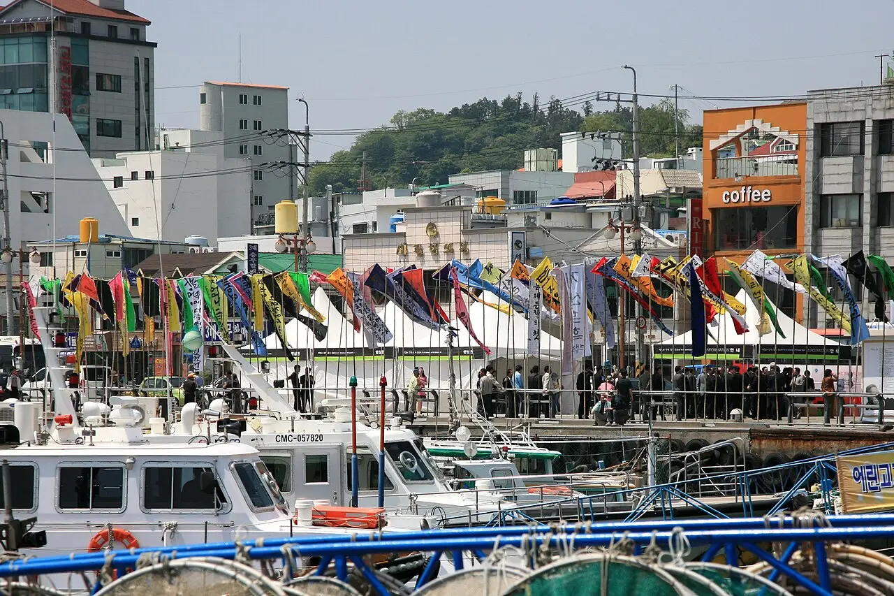 Porto de Tongyeong com barcos brancos em primeiro plano, bandeirolas coloridas e tendas ao longo do cais e prédios da cidade atrás.