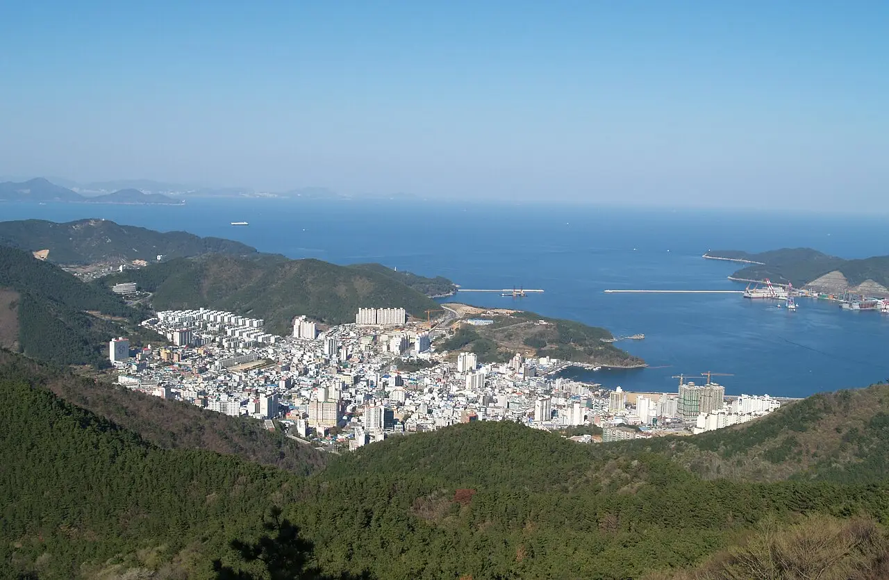 Vista do alto de Okpo, na ilha de Geoje, com a cidade de prédios claros junto a uma baía, colinas cobertas de pinheiros e estaleiros à direita.