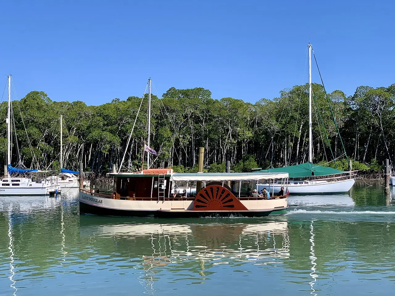 Barco de passeio de roda de pás Lady Douglas navegando em Port Douglas, entre veleiros ancorados e uma margem de manguezal.