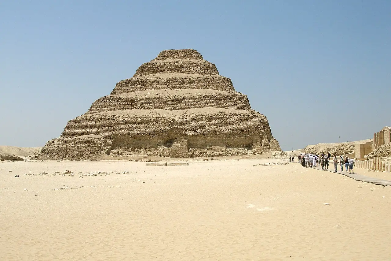 Pirâmide de degraus de Djoser em Saqqara, erguida sobre areia clara, com um grupo de visitantes caminhando por uma passarela à direita.