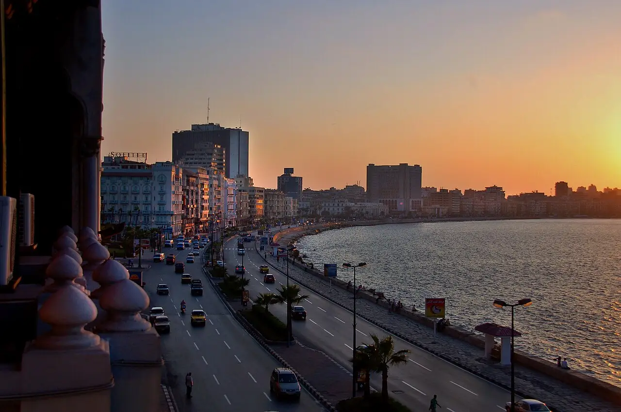 Avenida à beira-mar de Alexandria ao pôr do sol, com carros na pista, prédios ao longo da costa e o mar refletindo a luz alaranjada.