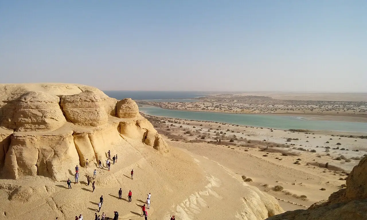 Visitantes sobre uma formação de arenito claro em Wadi al-Rayan, com um lago de água turquesa e o deserto se estendendo ao fundo.