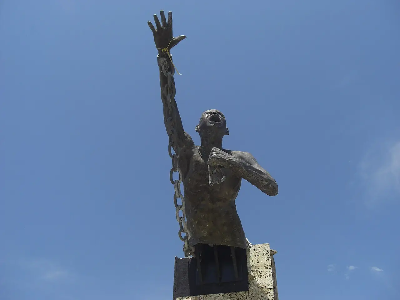 Estátua de bronze de um homem com o braço erguido e correntes, contra o céu azul.