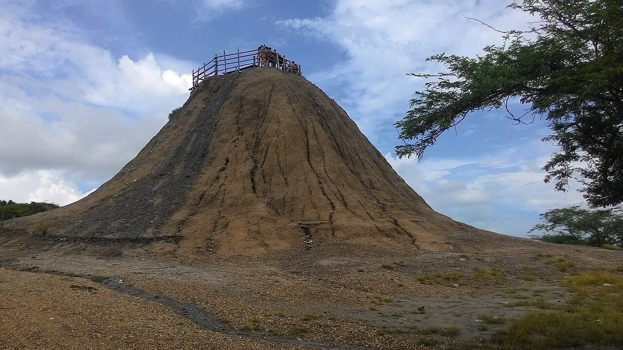 Cone de lama seca do Volcán del Totumo, com uma plataforma de madeira e um grupo de pessoas no topo, sob céu com nuvens.