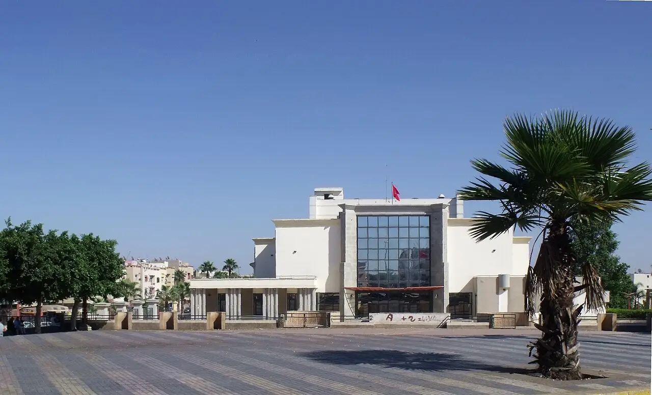 Teatro branco de fachada de vidro numa praça larga, com uma palmeira à direita.