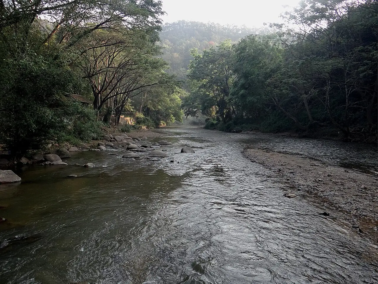 Um rio raso entre margens de pedra e mata fechada, com morros cobertos de árvore ao fundo.
