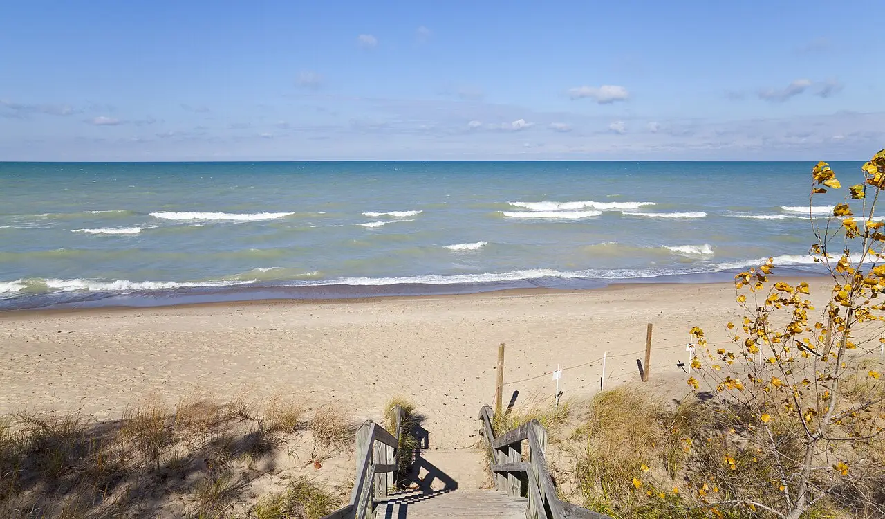 Escada de madeira descendo por dunas até a praia de areia do Indiana Dunes, com ondas do lago e o horizonte de água ao fundo.