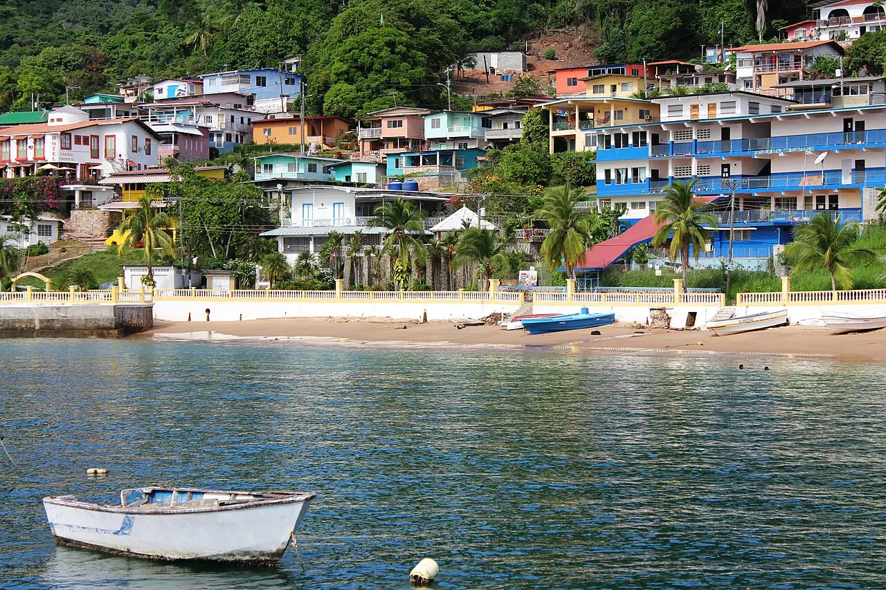 Praia e casas coloridas na encosta da Ilha Taboga, vistas da água, com um barco branco de remos em primeiro plano e mata ao fundo.