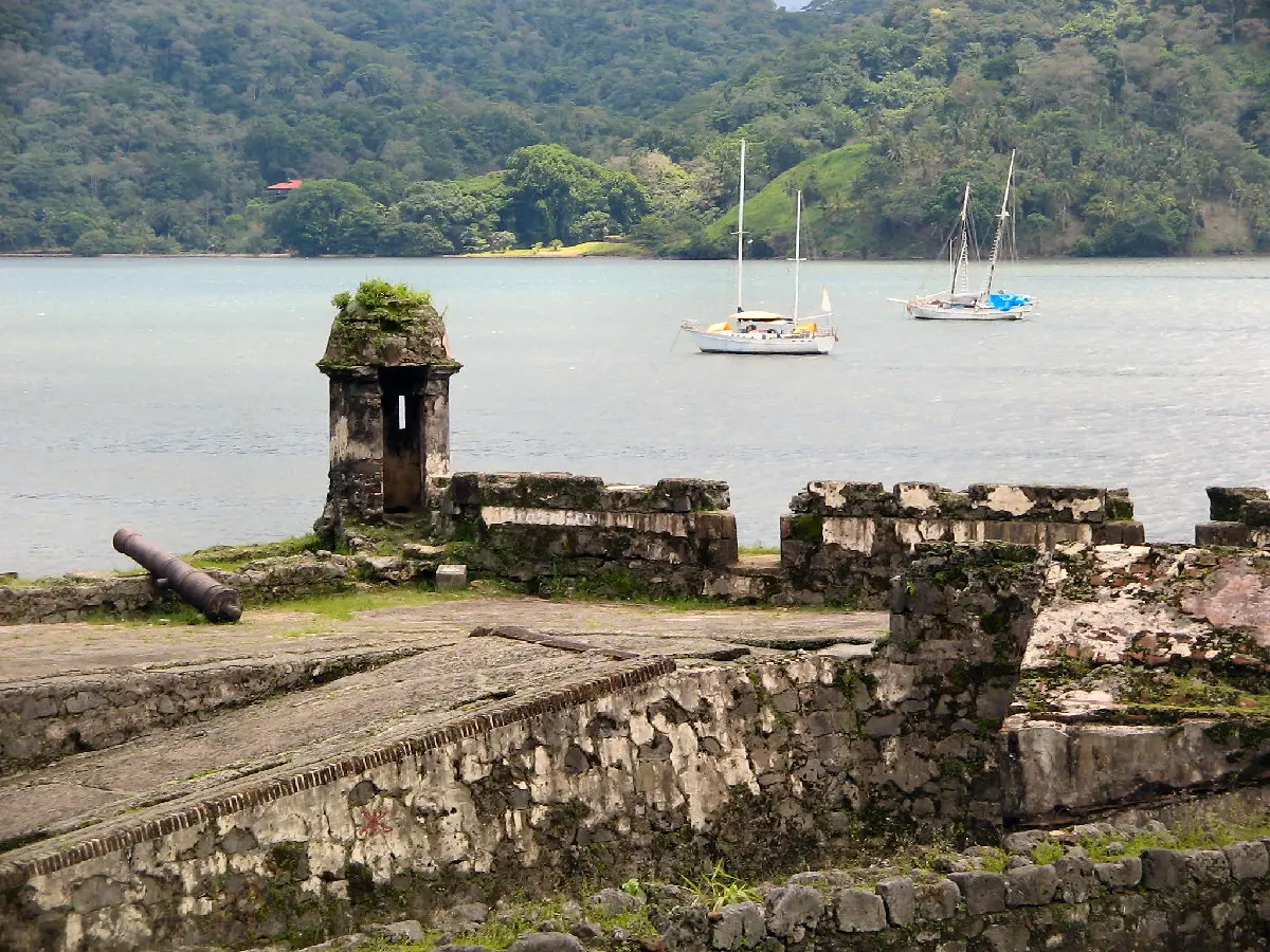 Muralha de pedra coberta de musgo, guarita e um canhão nas ruínas de Portobelo, com dois veleiros ancorados na baía e colinas de mata ao fundo.