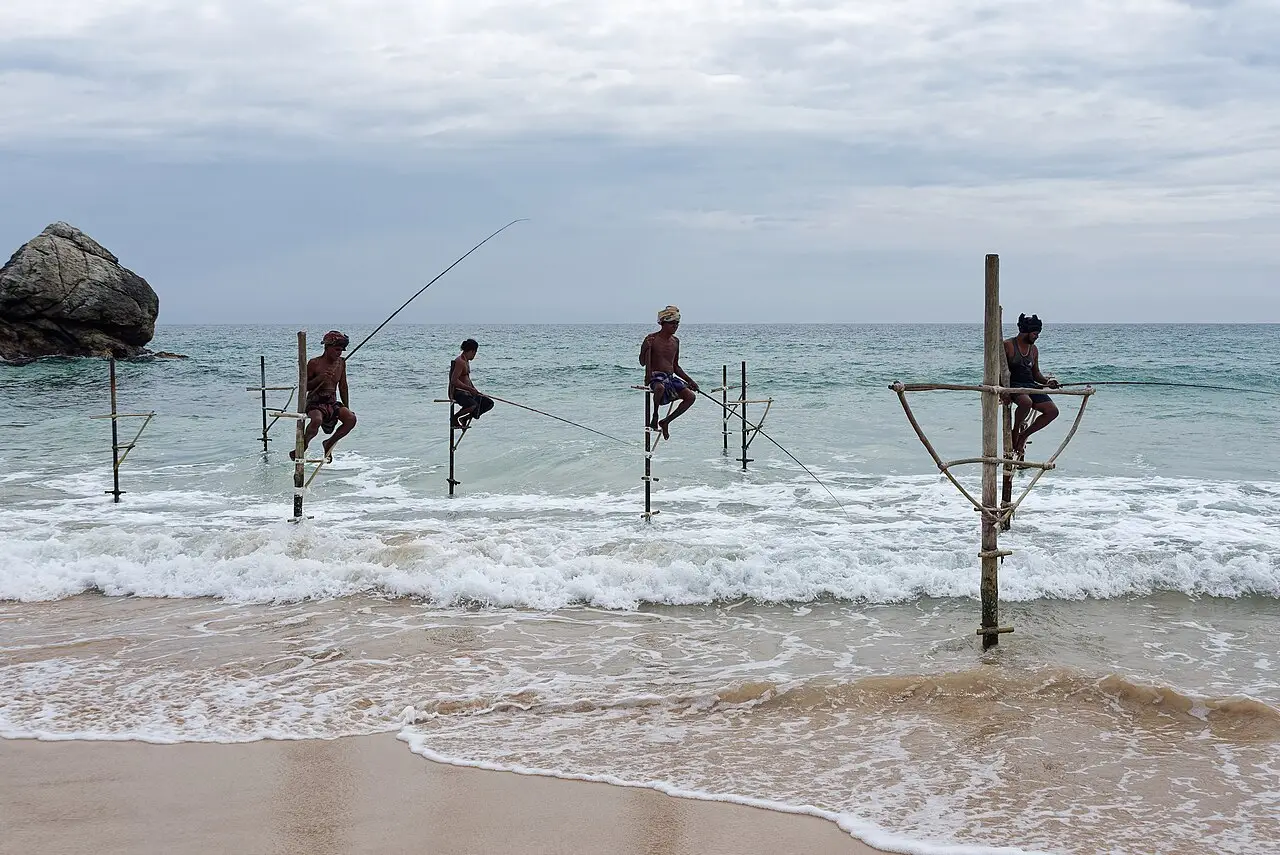 Pescadores sentados em estacas fincadas na arrebentação, com varas de pesca, perto de Galle, com uma grande rocha à esquerda e mar aberto ao fundo.