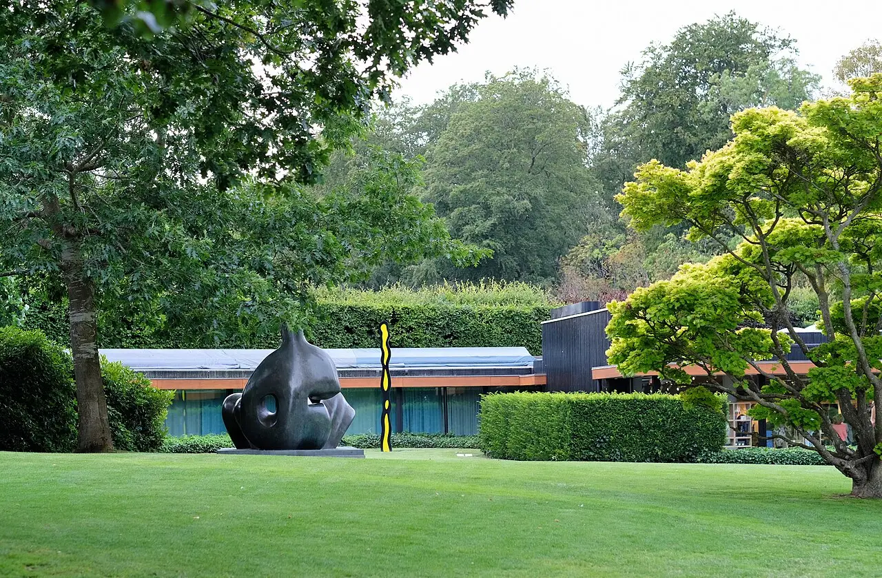 Jardim do Museu Louisiana com gramado, uma escultura de bronze escuro e uma coluna amarela e preta em frente ao prédio baixo do museu entre árvores.
