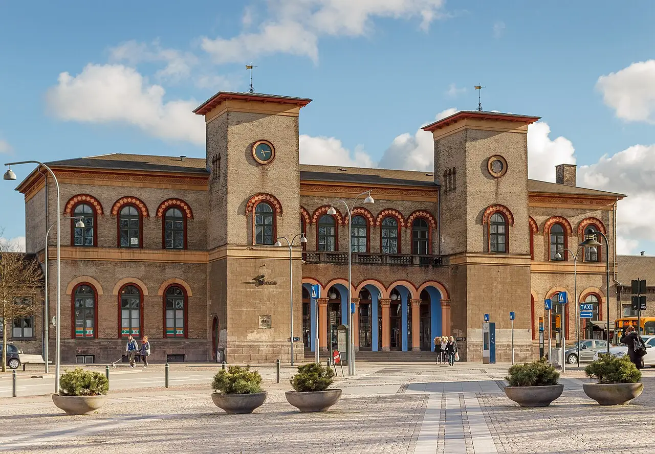 Edifício de tijolos da estação de Roskilde, com duas torres de relógio, arcos em vermelho e azul na entrada e vasos de plantas na praça.