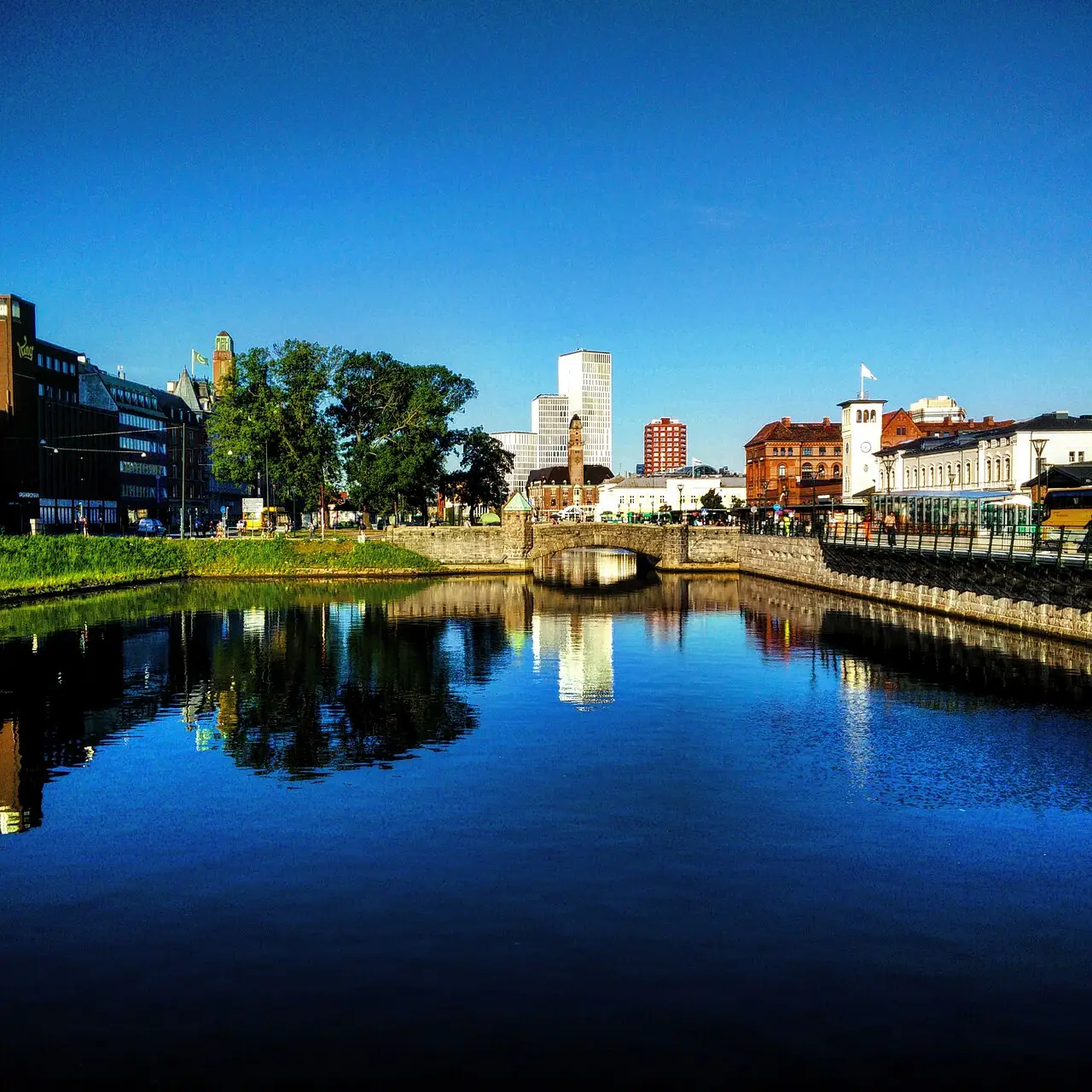 Um canal reflete prédios e uma ponte, sob céu azul.