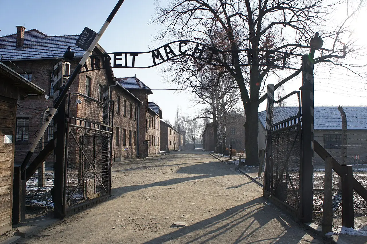 Portão de entrada de Auschwitz I com o letreiro de ferro em alemão sobre a passagem, barracões de tijolo ao fundo e cercas de arame farpado.