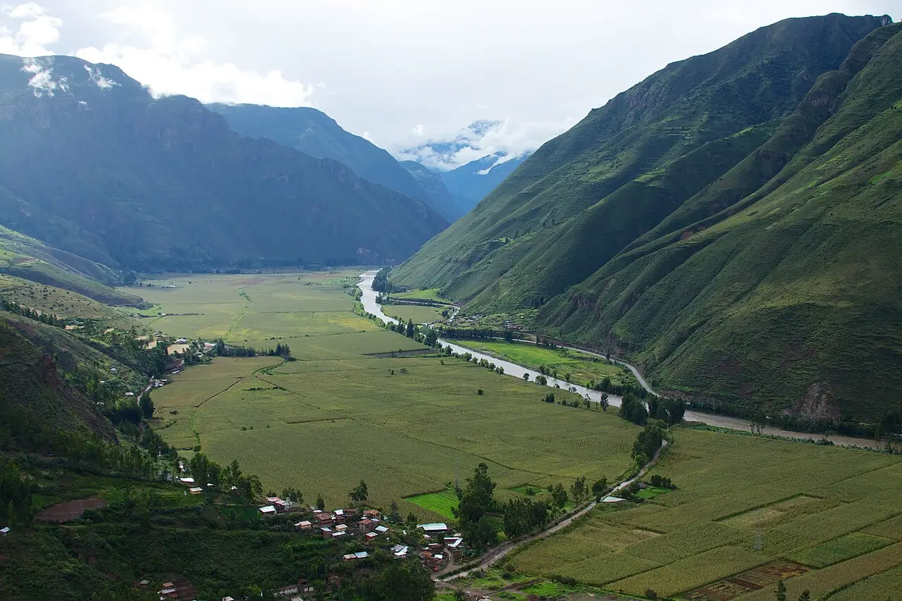 Vista do alto do Vale Sagrado, com um rio sinuoso no fundo plano coberto de campos, um povoado de casas pequenas e encostas verdes íngremes.