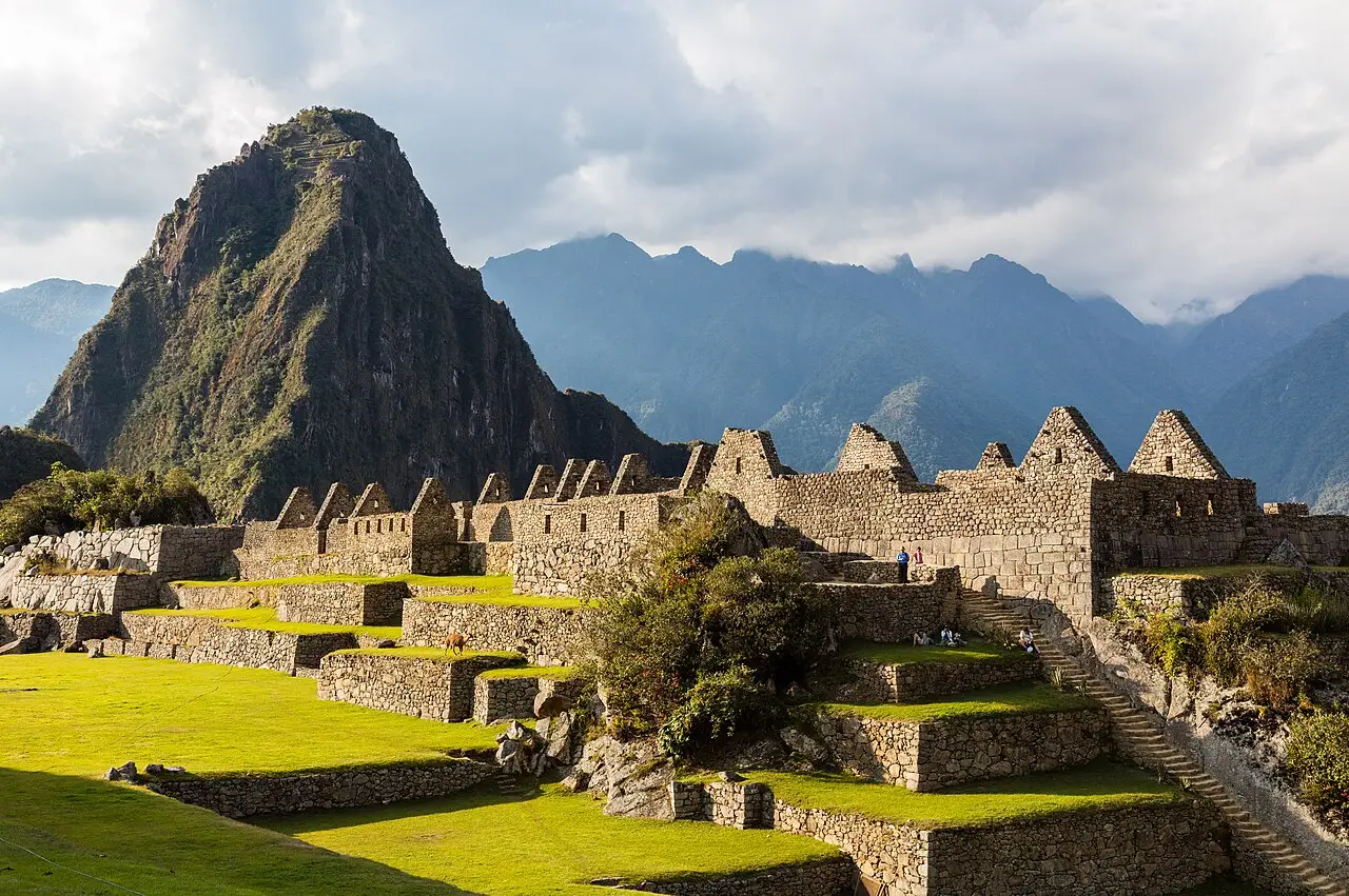 Construções de pedra e terraços gramados de Machu Picchu, com uma montanha pontiaguda coberta de mata à esquerda e cordilheira envolta em nuvens ao fundo.