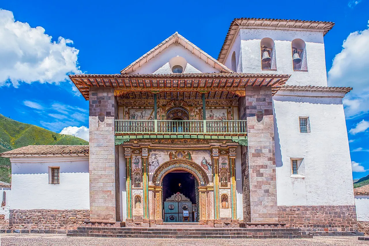 Fachada branca da igreja de Andahuaylillas, com portal pintado, balcão de madeira e torre com dois sinos.