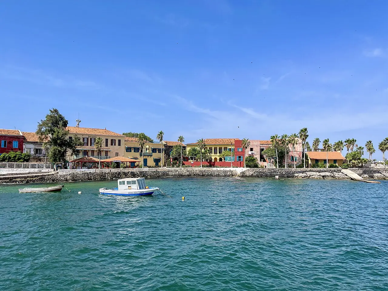 Orla da Île de Gorée vista do mar, com casas de fachadas coloridas e telhados de telha, palmeiras, um muro de pedra e barcos pequenos na água.