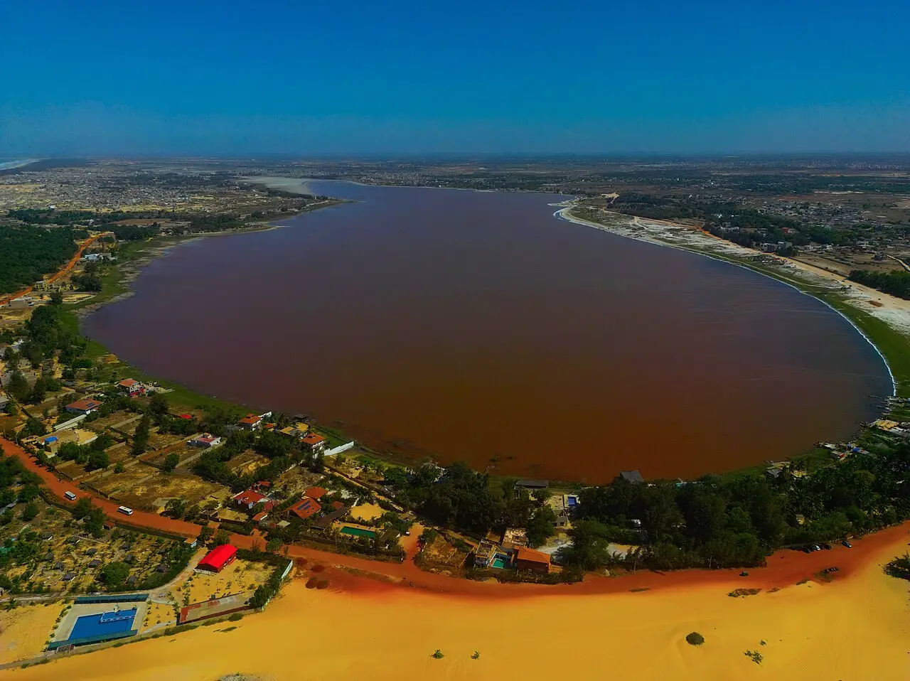 Vista aérea do Lac Rose, lago de água avermelhada cercado por casas e vegetação, com uma faixa de dunas de areia amarela em primeiro plano.