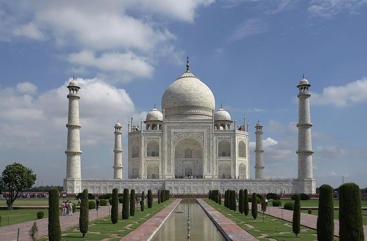 Taj Mahal visto de frente, com a cúpula de mármore branco, quatro minaretes, um canal de água central e jardins com ciprestes aparados.