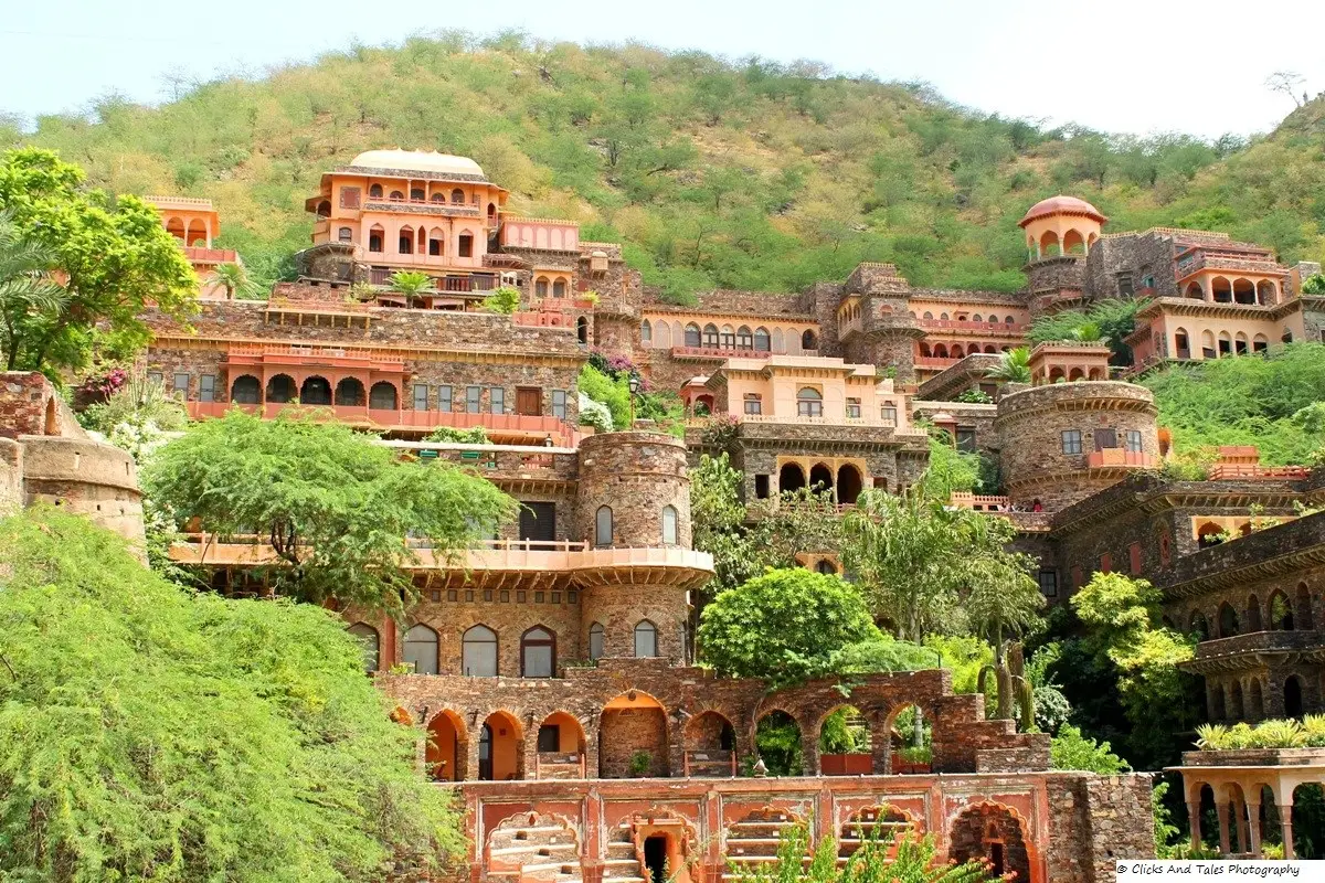 Fort Palace de Neemrana em terraços na encosta de um morro verde, com fachadas de pedra e tom rosado, arcos, varandas e cúpulas pequenas.