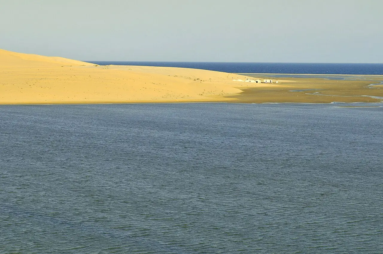 Duna de areia dourada chegando à água de uma enseada em Khor Al Adaid, com o mar aberto e um pequeno grupo de construções ao fundo.