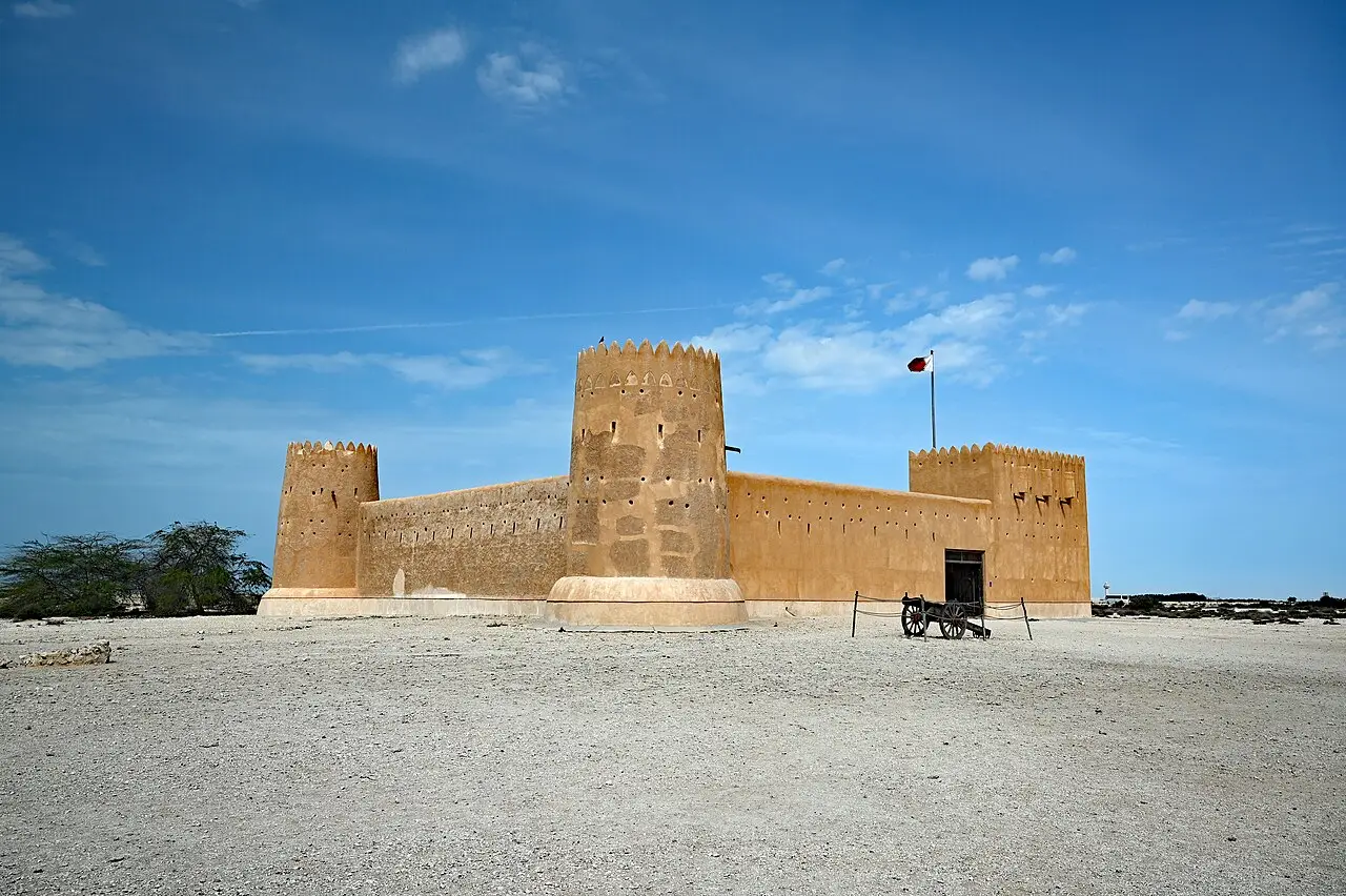 Forte de Al Zubarah, de paredes de tom areia com torres redondas e quadradas de ameias, uma bandeira e um canhão de rodas na frente.