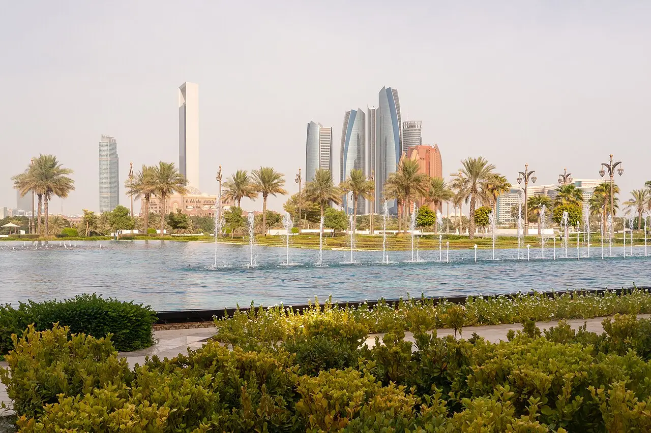 Arranha-céus do centro de Abu Dhabi vistos dos jardins do Qasr Al Watan, com fontes num espelho d’água, palmeiras e arbustos em primeiro plano.