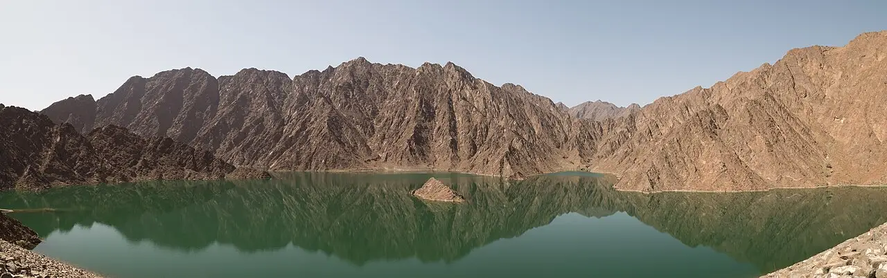 Panorama da represa de Hatta, com água verde-escura refletindo montanhas rochosas de tons marrons e uma pequena ilhota de pedra no meio.
