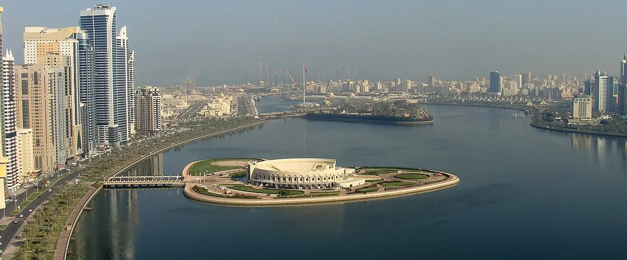 Vista aérea do Anfiteatro Al Majaz em Sharjah, numa ilha artificial com jardins ligada à margem por uma ponte, com prédios altos ao longo da laguna.