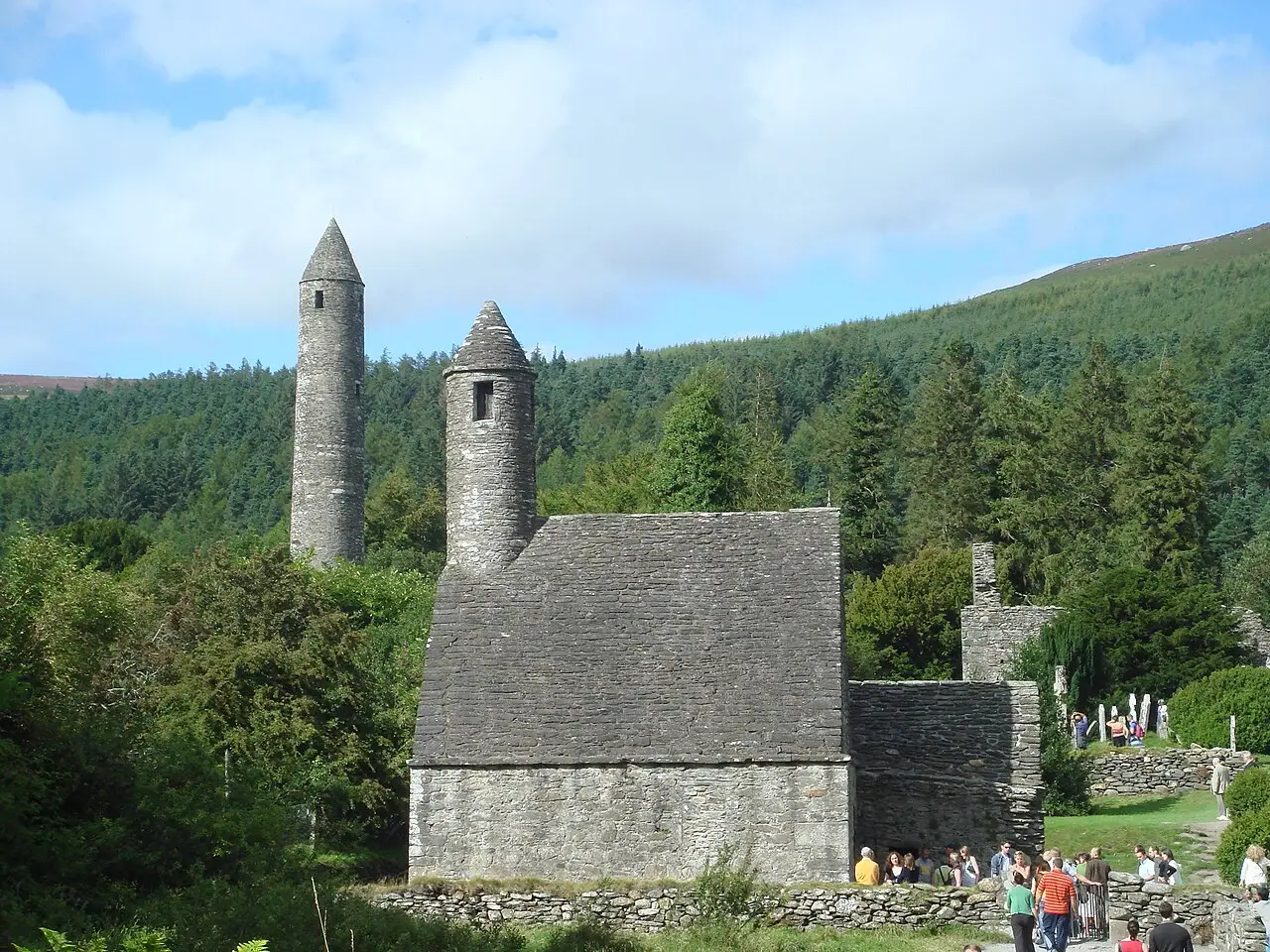 Ruínas do mosteiro de Glendalough, com a capela de telhado de pedra e uma torre redonda alta entre árvores, visitantes e colina de floresta ao fundo.