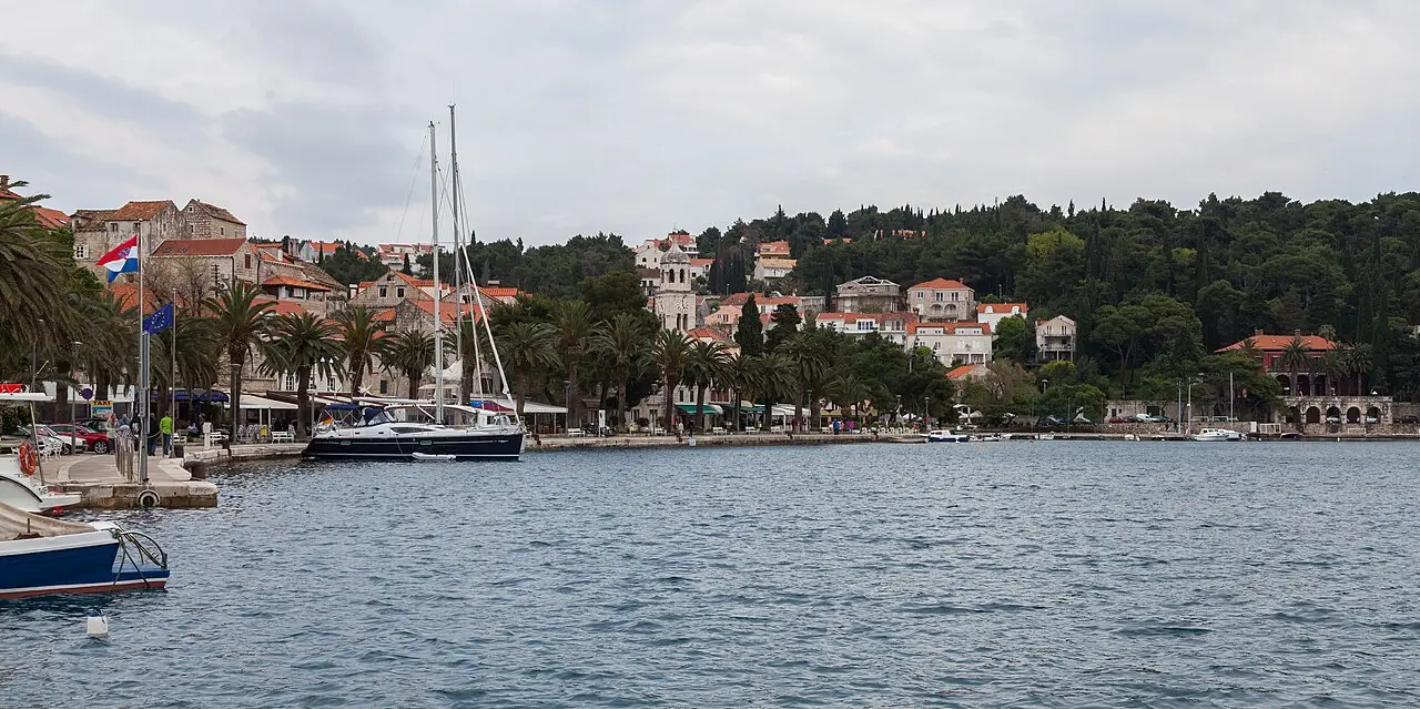 Orla de Cavtat vista da água, com veleiros e barcos atracados, palmeiras, casas de telhado vermelho, bandeira croata e colina de pinheiros ao fundo.