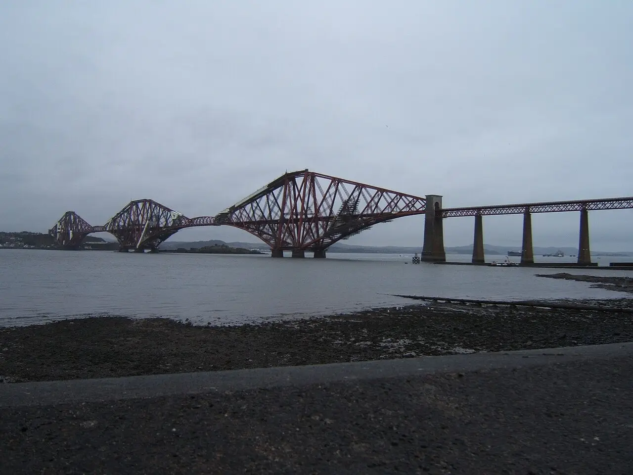Ponte ferroviária do Forth, de estrutura de aço vermelha com vãos em cantiléver, vista da margem de South Queensferry sob céu cinzento.
