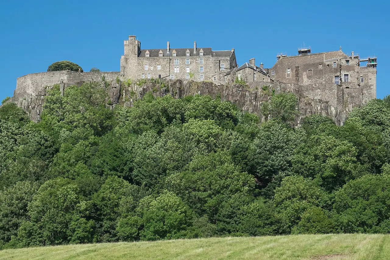 Castelo de Stirling no alto de um penhasco rochoso, com muralhas e edifícios de pedra acima de uma encosta coberta de árvores, sob céu azul.