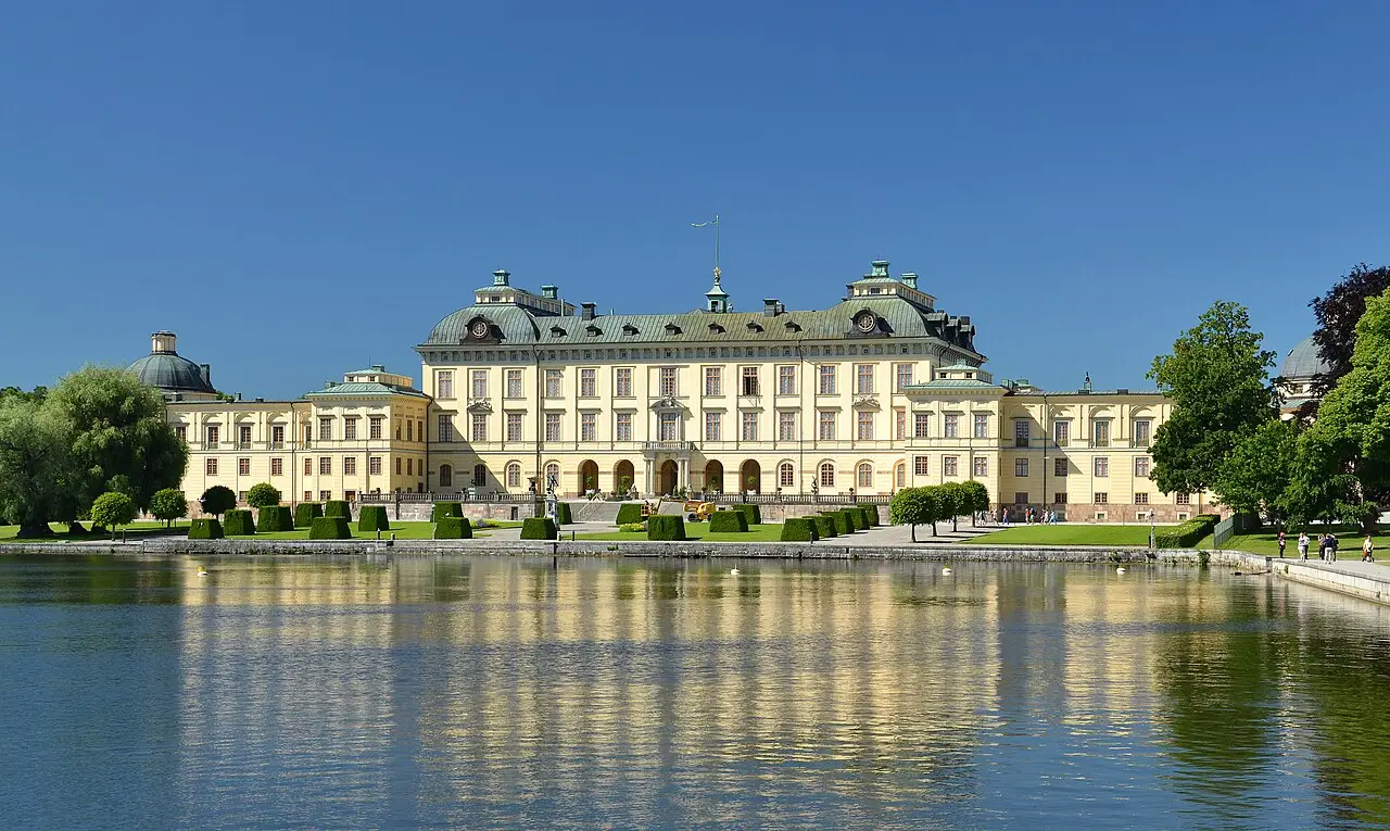 Façade jaune du palais de Drottningholm avec un toit de cuivre verdi, un jardin d’arbustes taillés et le reflet du bâtiment dans l’eau au premier plan.