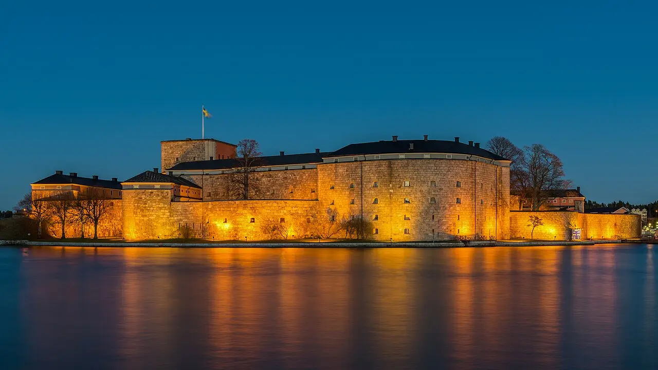 Fortaleza de Vaxholm iluminada ao anoitecer, com muros grossos de pedra arredondados e uma bandeira sueca, refletida na água calma.